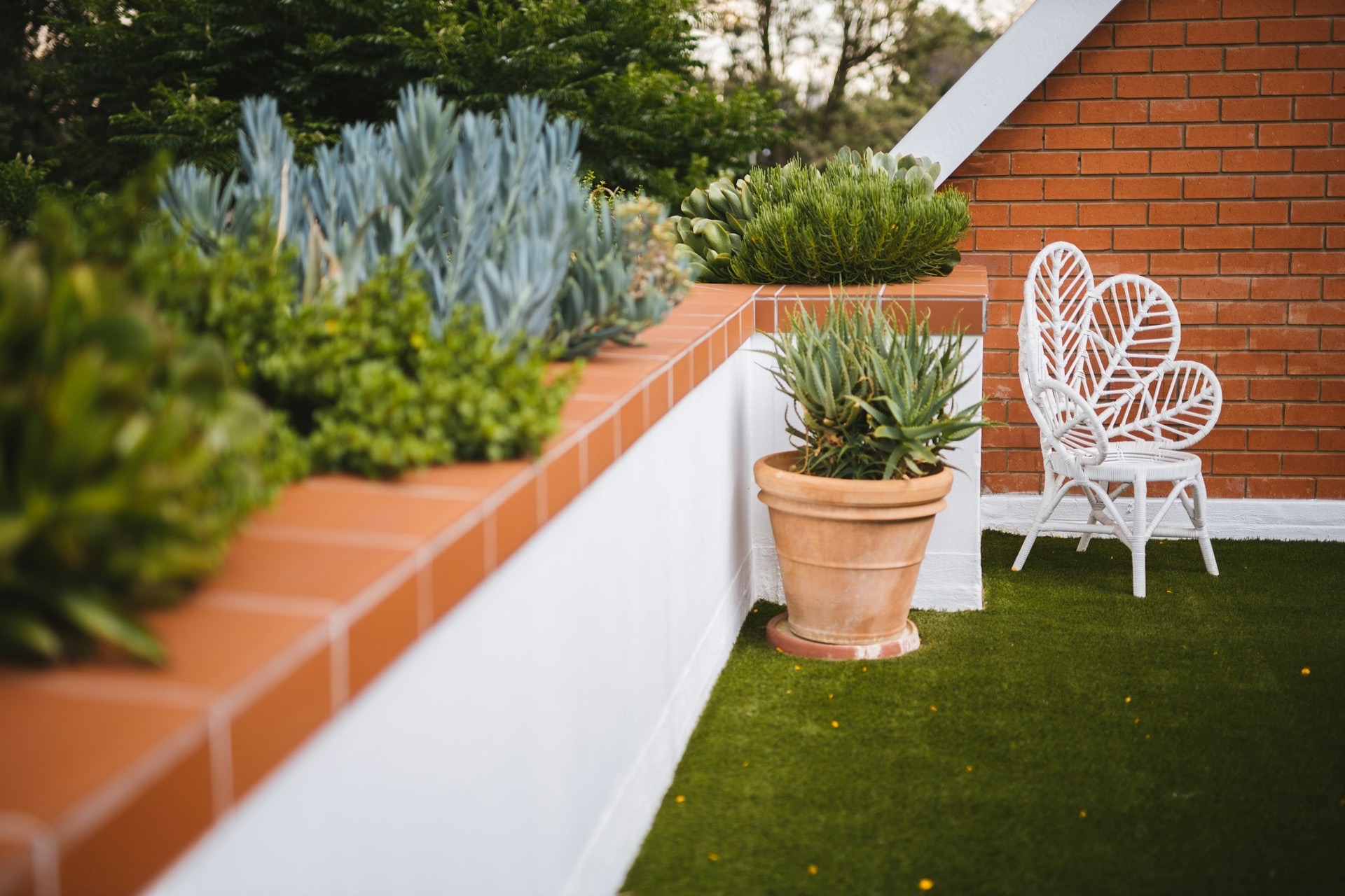 Small courtyard corner with a white cane chair on artificial grass beside a brick wall and terracotta planters